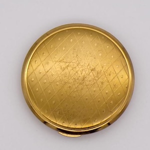 Op de afbeelding: Een ronde, goudkleurige compactpoederdoos met een ruitpatroon dat in het oppervlak is geëtst. De compactpoederdoos heeft een scharnierende opening en vertoont lichte gebruikssporen, wat duidt op de leeftijd. Het ontwerp is eenvoudig en elegant.