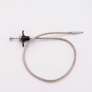 May include: A camera shutter release cable featuring a flexible, braided silver-toned metal cord, a black plastic plunger handle with a chrome-plated button, and a threaded metal tip for attachment to a camera body for remote shutter operation.