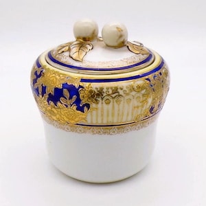 Może przedstawiać: Biała porcelanowa cukiernica z pokrywką. Miseczka posiada niebieski pas ze złotymi wzorami kwiatowymi i złotymi zdobieniami. Pokrywka ma dwa okrągłe gałki i złote akcenty liści. Cukiernica to przedmiot dekoracyjny.