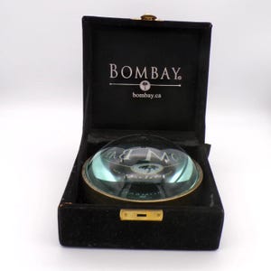 May include: A clear glass paperweight with a gold-coloured rim, in a black velvet box. The paperweight reflects the text "BOMBAY" and "bombay.ca". The box has a gold-coloured clasp.