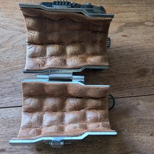 May include: Two brown leather wrist cuffs with silver metal buckles and clasps. The cuffs are connected by a silver metal hinge.