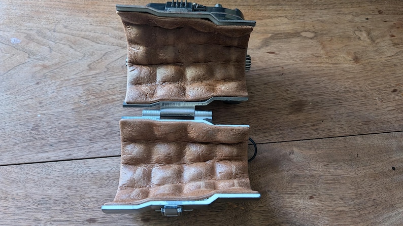 May include: Two brown leather wrist cuffs with silver metal buckles and clasps. The cuffs are connected by a silver metal hinge.