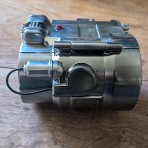 May include: A silver-colored metal object with a black wire attached. The object has a small red button and a hinge on the side. The object is on a wooden surface.