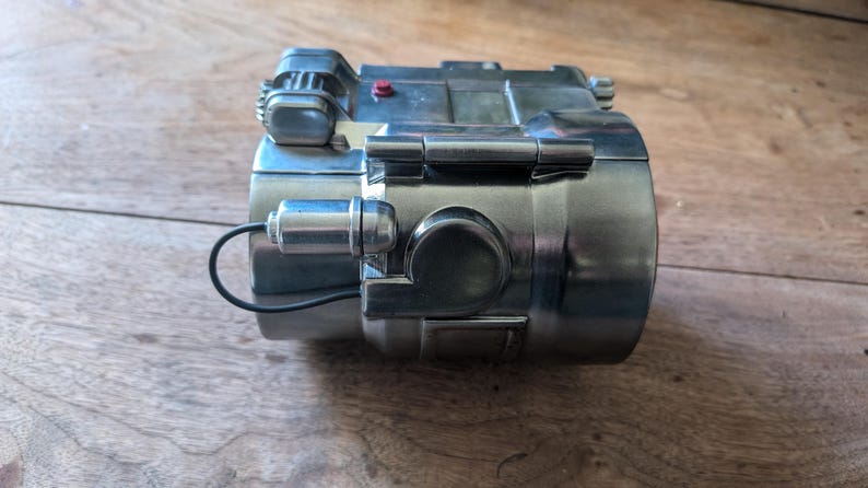 May include: A silver-colored metal object with a black wire attached. The object has a small red button and a hinge on the side. The object is on a wooden surface.