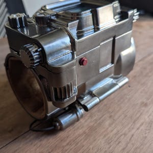 May include: A silver metal device with a round, cylindrical base and a complex, multi-faceted top. The device has a red button on the side and several gears and levers visible. The device appears to be a mechanical or electronic device.
