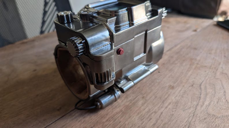 May include: A silver metal device with a round, cylindrical base and a complex, multi-faceted top. The device has a red button on the side and several gears and levers visible. The device appears to be a mechanical or electronic device.