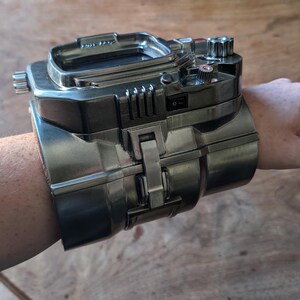 May include: A silver metal wristwatch with a digital display and multiple buttons and dials. The watch is designed to look like a futuristic gadget.