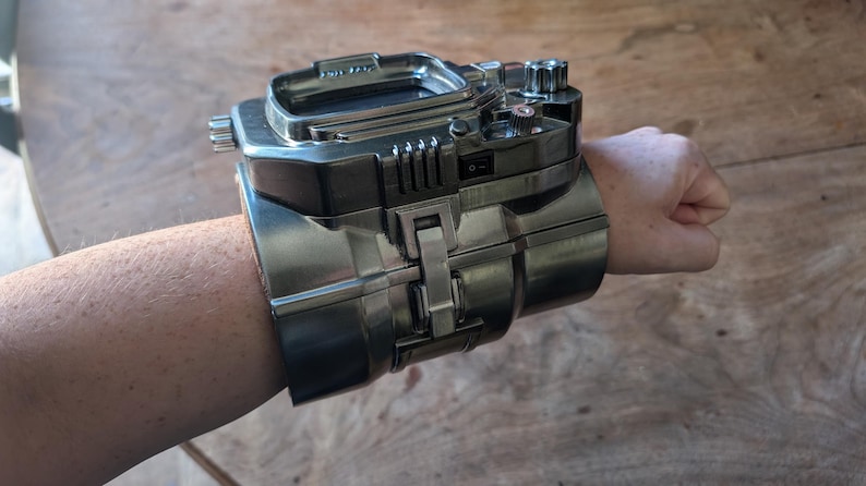 May include: A silver metal wristwatch with a digital display and multiple buttons and dials. The watch is designed to look like a futuristic gadget.