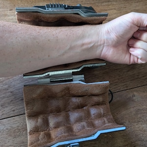 May include: Two brown leather wristbands with a metal hinge and a silver metal gear mechanism. The wristbands are open and laid flat on a wooden surface.