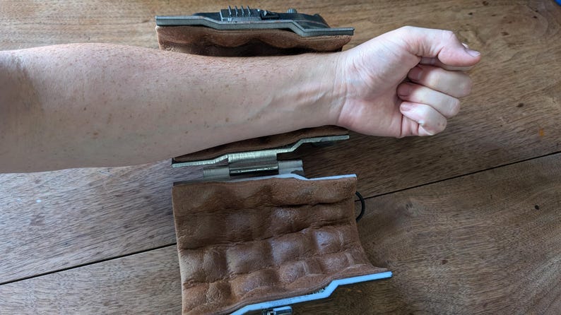 May include: Two brown leather wristbands with a metal hinge and a silver metal gear mechanism. The wristbands are open and laid flat on a wooden surface.