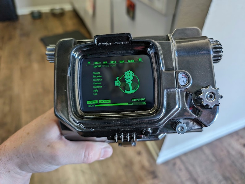 May include: A Pip-Boy device with a green screen displaying a character's stats, including Strength, Perception, Endurance, Charisma, Intelligence, Agility, Luck, and Special Perks. The screen also shows a thumbs up icon and the text 'SPECIAL PERKS'. The device has a silver metal casing with a black dial and a gear on the side.