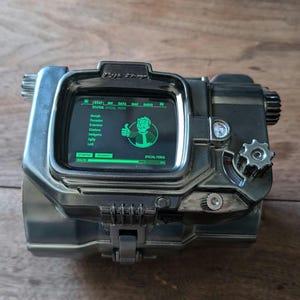 May include: A black and silver Pip-Boy device with a green screen displaying text and a cartoon character. The screen reads "START" and "SPECIAL" and shows a character with a thumbs up.