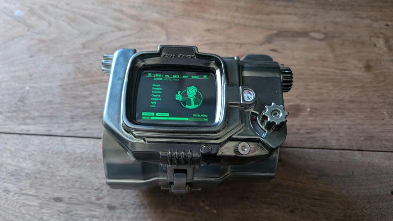 May include: A black and silver Pip-Boy device with a green screen displaying text and a cartoon character. The screen reads "START" and "SPECIAL" and shows a character with a thumbs up.
