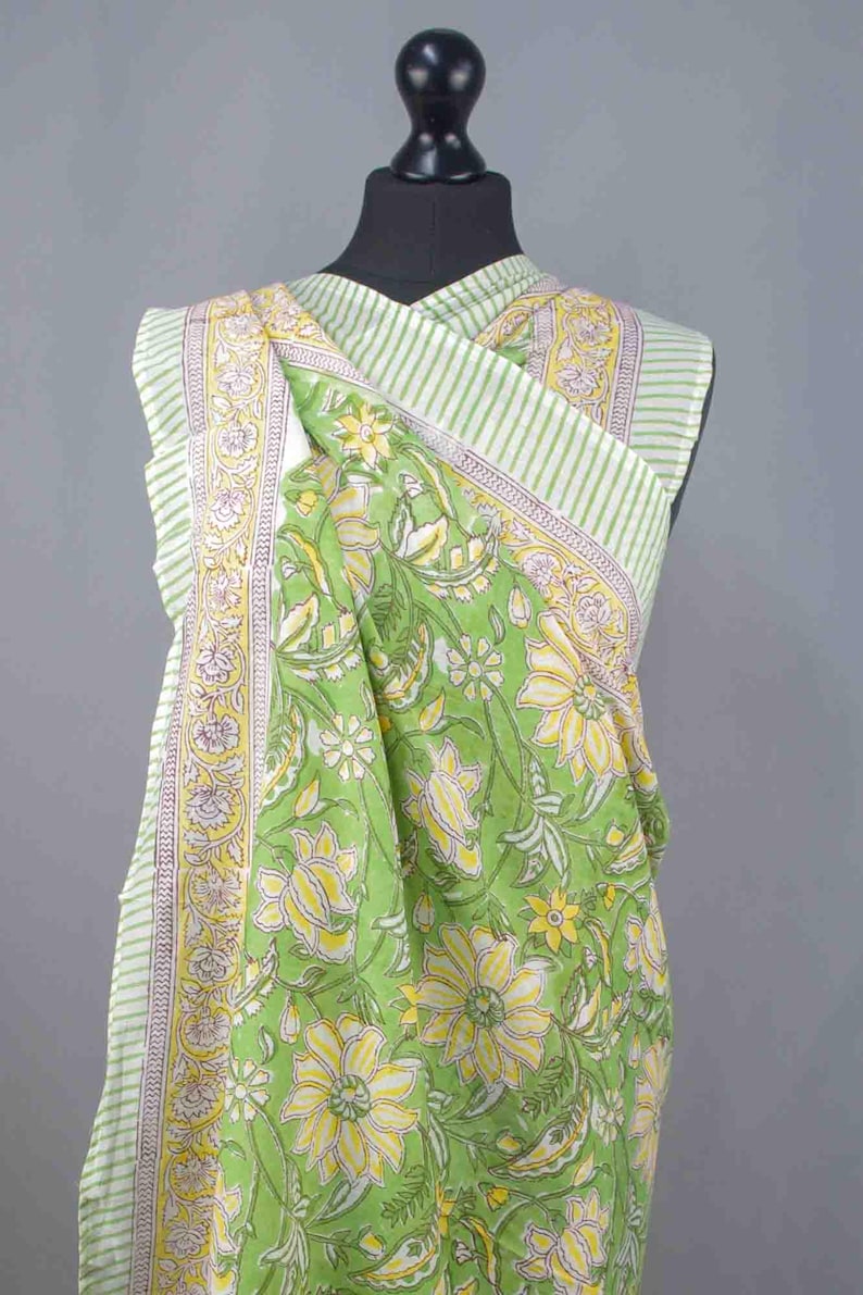Puede incluir: Textil verde con estampado floral drapeado sobre un maniqu&iacute;. La tela presenta grandes flores amarillas y hojas verdes sobre un fondo verde claro. El borde tiene un patr&oacute;n de rayas con acentos amarillos y morados.