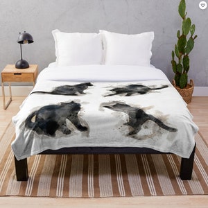 May include: A white throw blanket with a watercolor design of four black cats in different poses. The cats are painted in a loose, abstract style, with soft edges and subtle shading.