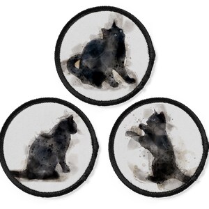 Könnte beinhalten: Drei Aquarell-Illustrationen von schwarzen Katzen auf weißen runden Patches mit schwarzen Nähten. Die Katzen sind in verschiedenen Posen, eine sitzt, eine steht und eine springt.