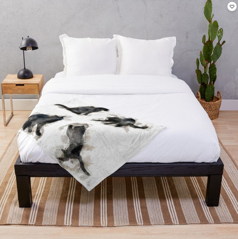 May include: A white throw blanket with a watercolor design of four black cats in various poses. The blanket is on a bed with a black frame and a brown and white striped rug.