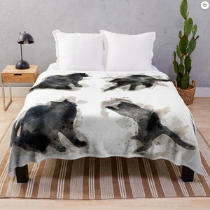 May include: A white throw blanket with a watercolor design of four black cats in different poses. The cats are painted in a loose, abstract style with splashes of gray and brown.