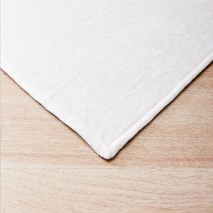 May include: Close-up of the edge of a white, soft, plush blanket. The blanket is folded and the edge is visible against a light brown wooden surface.