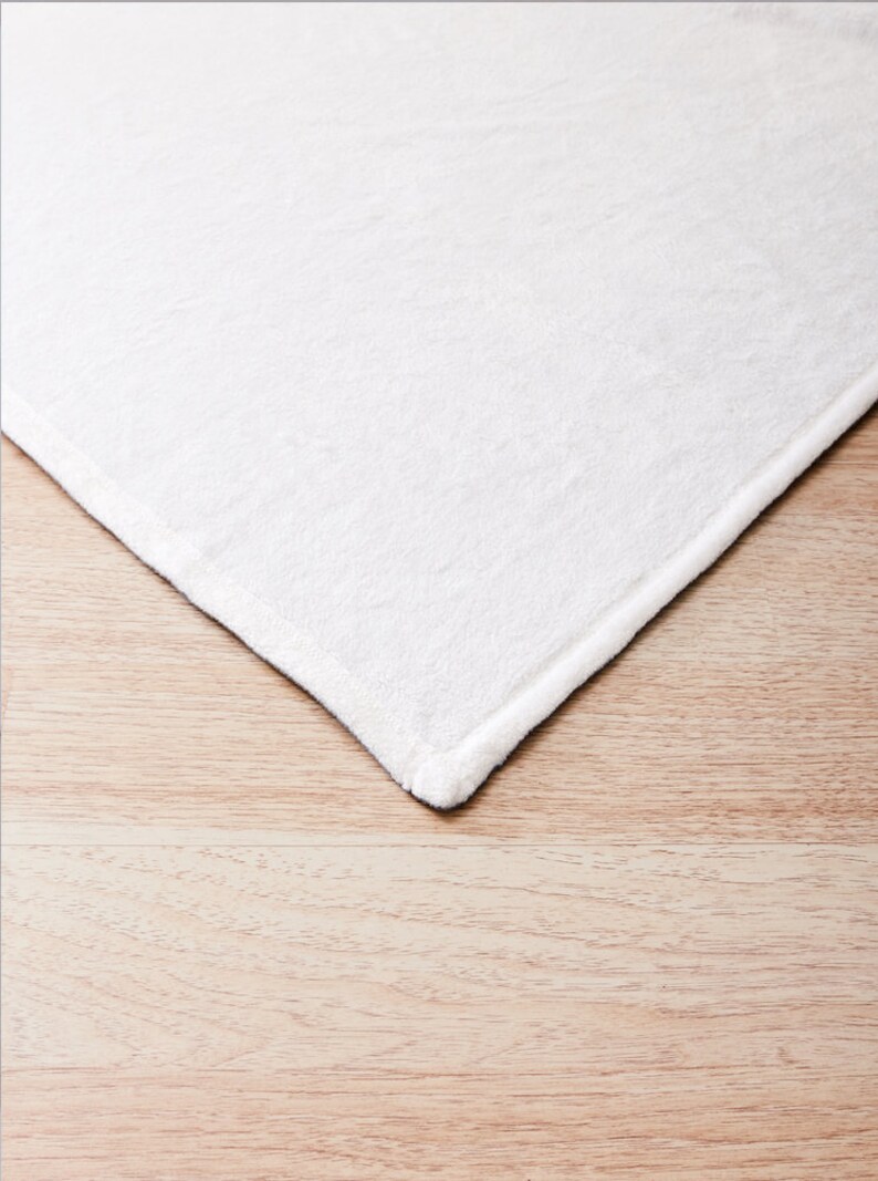 May include: Close-up of the edge of a white, soft, plush blanket. The blanket is folded and the edge is visible against a light brown wooden surface.