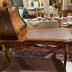 Puede incluir: Un escritorio de madera vintage con una tapa de cuero y un gabinete incorporado con cajones. El escritorio tiene tallas ornamentadas y detalles dorados. El escritorio est&aacute; sobre una alfombra con un patr&oacute;n floral.