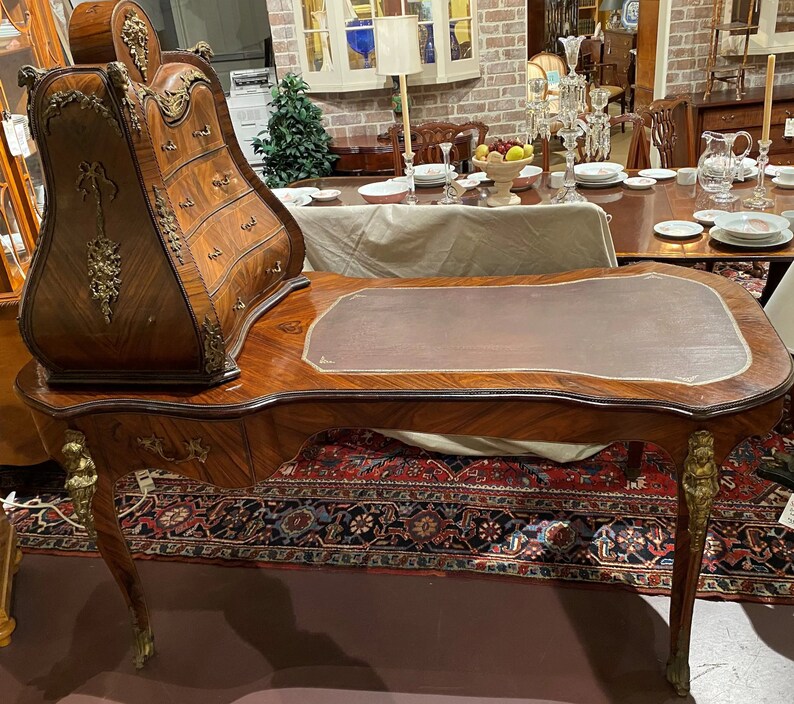 Puede incluir: Un escritorio de madera vintage con una tapa de cuero y un gabinete incorporado con cajones. El escritorio tiene tallas ornamentadas y detalles dorados. El escritorio est&aacute; sobre una alfombra con un patr&oacute;n floral.