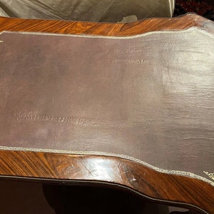 Puede incluir: Un escritorio de madera marr&oacute;n oscuro con una tapa de cuero. La tapa de cuero tiene un borde dorado decorativo y un borde ligeramente curvado. El escritorio tiene una rica veta de madera.
