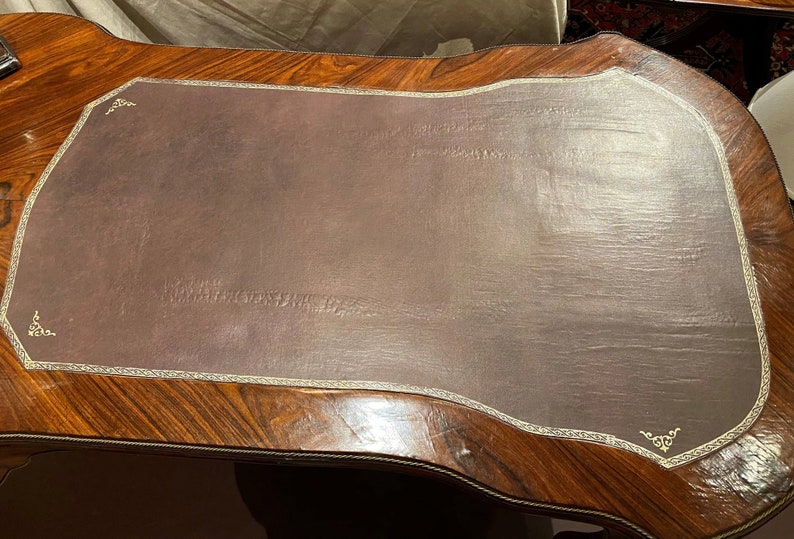 Puede incluir: Un escritorio de madera marr&oacute;n oscuro con una tapa de cuero. La tapa de cuero tiene un borde dorado decorativo y un borde ligeramente curvado. El escritorio tiene una rica veta de madera.
