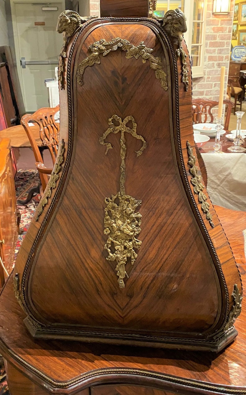 Puede incluir: Un gabinete de madera con una parte superior curva y adornada y detalles intrincados de lat&oacute;n. El gabinete est&aacute; hecho de madera oscura con un patr&oacute;n de chevron y presenta un lazo de lat&oacute;n decorativo y un dise&ntilde;o floral.