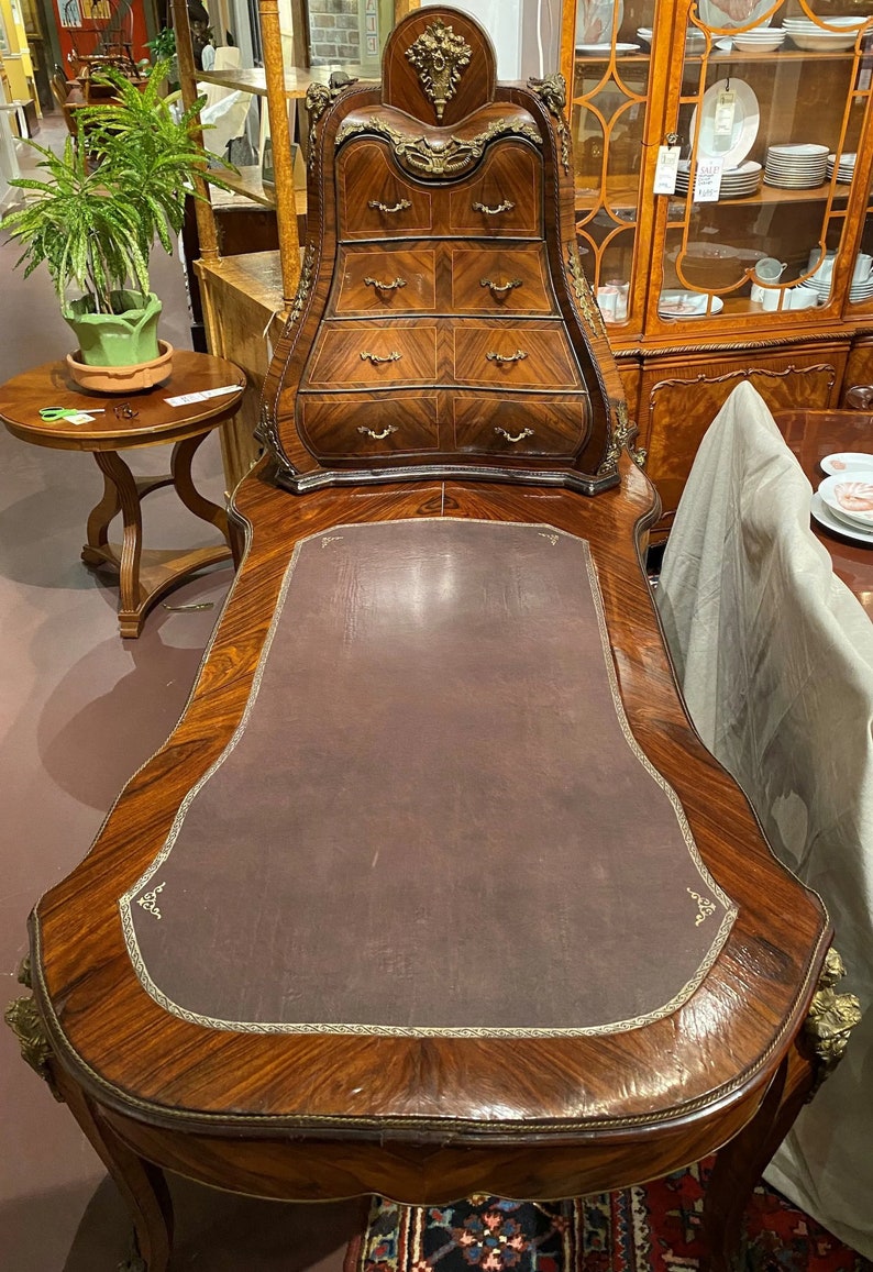 Puede incluir: Un escritorio de madera vintage con una tapa de cuero marr&oacute;n y una secci&oacute;n superior curvada y adornada con varios cajones. El escritorio tiene una rica veta de madera y detalles intrincados.