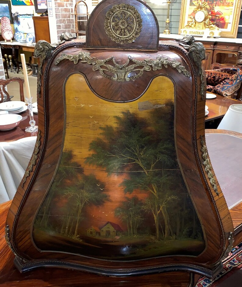 Puede incluir: Un gabinete de madera vintage con una escena pintada de un bosque con una peque&ntilde;a casa. El gabinete tiene adornos de lat&oacute;n ornamentados y una parte superior curva.