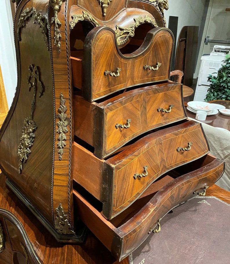 Puede incluir: Una c&oacute;moda antigua de madera con tallas intrincadas y detalles en lat&oacute;n. La c&oacute;moda tiene cuatro cajones con frentes curvos y tiradores ornamentados. Los cajones est&aacute;n abiertos, revelando la veta de la madera y el interior de la c&oacute;moda.