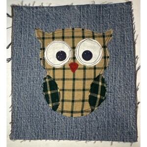 May include: A brown and green plaid owl with white eyes and a red beak is sewn onto a blue denim background.