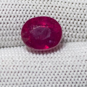 May include: A vivid, oval-cut ruby gemstone, set against a white, textured fabric. The ruby exhibits a deep red colour, with visible internal features. The setting is neutral, highlighting the gem's natural beauty.