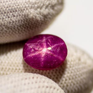 May include: A close-up of a round, polished, star ruby gemstone. The gemstone is a deep magenta color with a six-rayed star pattern visible on its surface. The ruby is held by a white glove, with a white background.