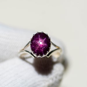 Op de afbeelding: Een zilveren ring met een grote, ovale ster-robijn edelsteen. De diep magenta steen vertoont een ster-effect. De band van de ring splitst zich aan weerszijden van de edelsteen in twee delicate strengen.