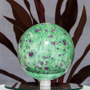 May include: A polished, spherical gemstone featuring a vibrant green base color with scattered, irregular deep magenta and dark red inclusions throughout its smooth, reflective surface, resting on a small, circular, light-colored stone base.