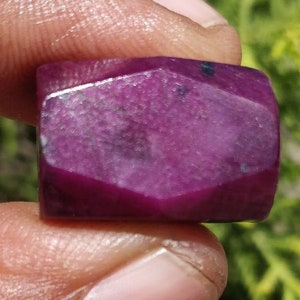 May include: A rectangular, faceted, deep magenta gemstone is held between fingers. The stone has a polished surface with subtle variations in color and small dark inclusions. The background is blurred green foliage.
