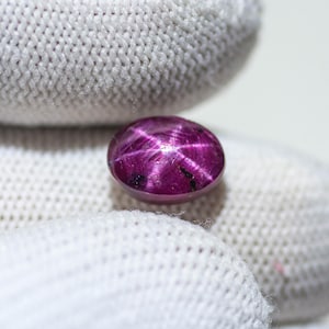 Puede incluir: Una piedra preciosa de rubí estrella ovalada y pulida. La gema es de color magenta intenso con un patrón de estrella de seis rayos en su superficie. El rubí está sobre un fondo blanco.
