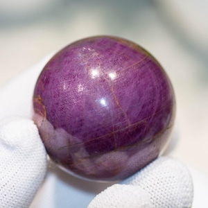 May include: A polished, spherical gemstone in shades of deep purple. The orb has a smooth surface with subtle internal veining and a slight sheen. The gemstone is held in a white gloved hand, highlighting its color and form.