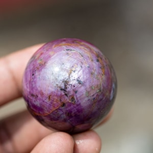 May include: A purple and silver sphere with a smooth, polished surface. The sphere is made of a natural gemstone, possibly ruby.
