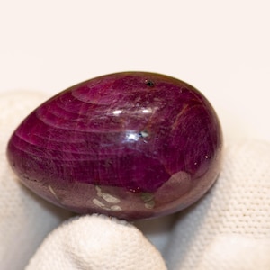 Pode incluir: Uma pedra preciosa polida, de forma oval, em um tom magenta profundo. A pedra tem uma superfície lisa e reflexiva com estrias internas visíveis. A gema é segurada em um tecido branco, destacando sua cor e forma. Esta é uma pedra preciosa.