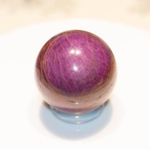 May include: A polished, spherical gemstone in shades of deep purple and reddish-brown. The orb appears to be resting on a clear, rounded base, with a white background. The gemstone's surface has a natural, textured appearance.