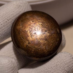 Könnte beinhalten: Ein polierter, oval geformter Edelstein mit metallisch bronzefarbener Farbe. Die Oberfläche hat einen reflektierenden Glanz mit sichtbaren Streifen. Der Stein wird in einem weißen Handschuh gehalten. Dies könnte ein dekorativer Artikel oder ein Sammlerstück sein.