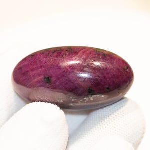 Op de afbeelding: Een gepolijste, ovaalvormige edelsteen in tinten diep magenta met donkere insluitsels. De steen is glad en reflecterend, tegen een witte achtergrond. De edelsteen wordt vastgehouden door een gehandschoende hand.