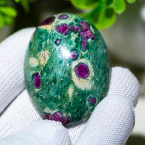 Könnte beinhalten: Ein ovaler Edelstein mit grüner Grundfarbe und leuchtend magentafarbenen Einschlüssen. Der Stein wird in einer weißen behandschuhten Hand gehalten, mit einer verschwommenen grünen Pflanze im Hintergrund. Der Edelstein hat eine polierte, glatte Oberfläche.