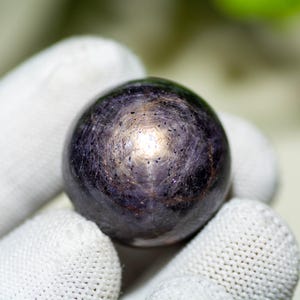 May include: A dark purple gemstone sphere with a star-like pattern. The polished surface reflects light, creating a shimmering effect. The gem is held by a gloved hand, highlighting its shape and color.