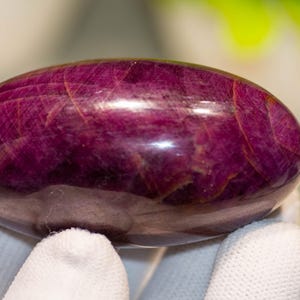 May include: A polished, oval-shaped gemstone in a deep magenta hue. The stone has a smooth, glossy surface with subtle internal veining. The gemstone is held by a gloved hand, highlighting its size and shape. This is a decorative item.