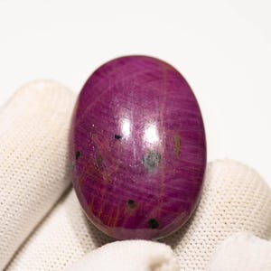 May include: A polished, oval ruby gemstone. The gem is a deep magenta colour with visible internal striations and small dark inclusions. The stone is held against a white background.
