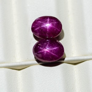 Peut inclure: Deux rubis étoilés ovales, d'un magenta profond. Chaque gemme présente un motif en étoile à six rayons. Les rubis sont posés sur un fond blanc, ce qui met en valeur leur couleur riche et leur effet optique unique. Ces pierres sont idéales pour la joaillerie.